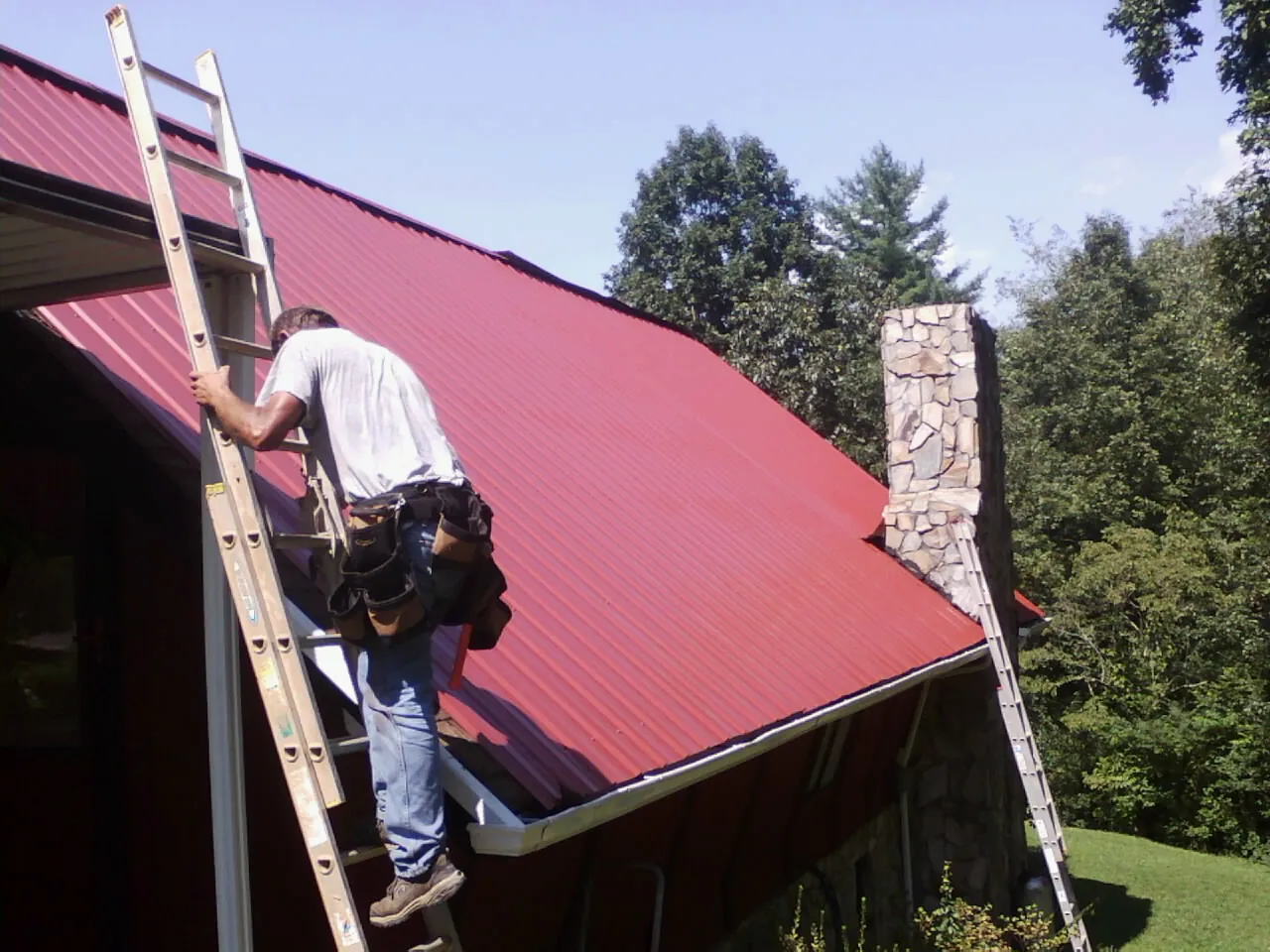 Roofer on a ladder beside a red metal roof and stone chimney — Soffit & Fascia Repair by Roofing Brothers in Wallowa